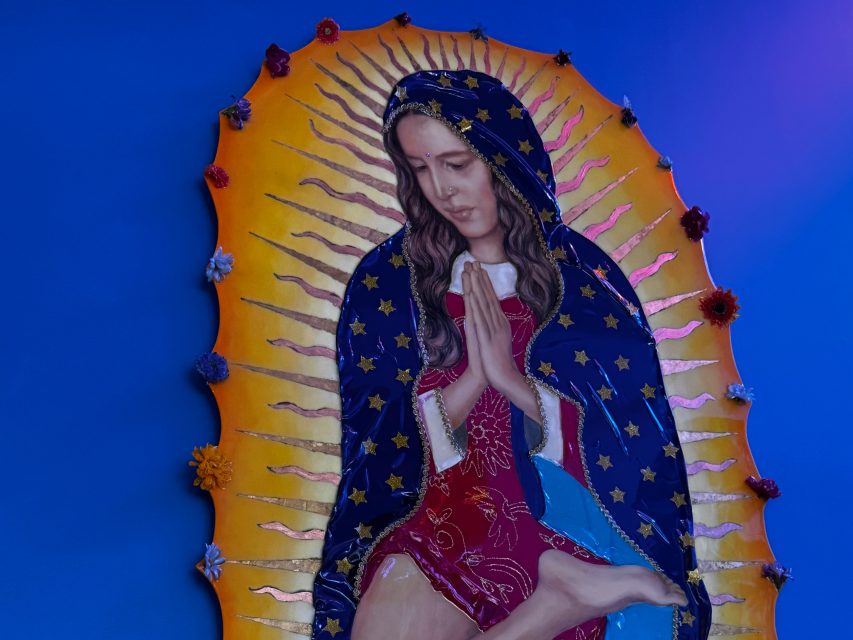 A statue of a woman in a red dress and blue cloak with gold stars is depicted praying, surrounded by a yellow radiance and flowers, against a blue background.