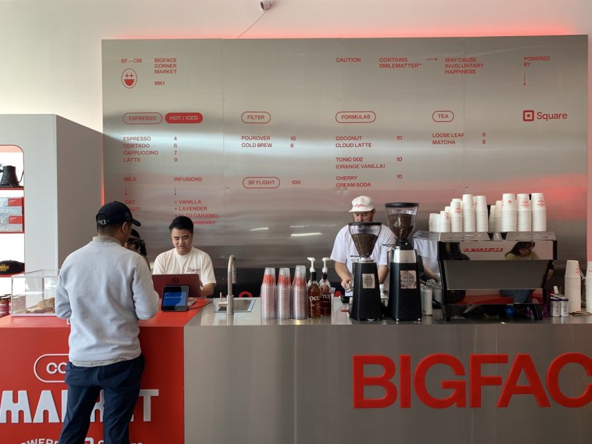 A man orders at a modern coffee counter with two staff members, espresso machines, a menu board, and stacks of cups in a minimalistic cafรฉ setting.