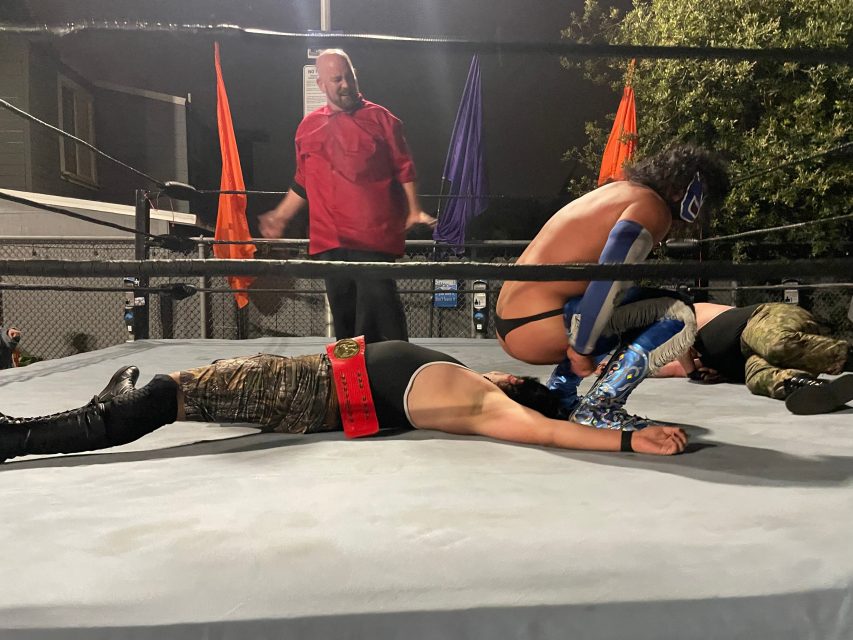 A wrestling match in progress: one masked wrestler pins an opponent on the mat while a referee observes and another wrestler lies motionless nearby.