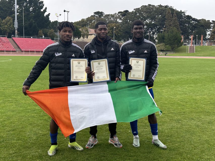 Three men standing on a grass field hold certificates and display the Irish flag. All are wearing black jackets and athletic attire. Stadium seating and trees are visible in the background.