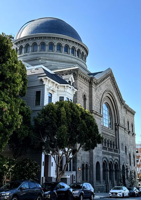 Stone building with a large domed roof and arched windows, partially obscured by trees, with parked cars along the street in front.