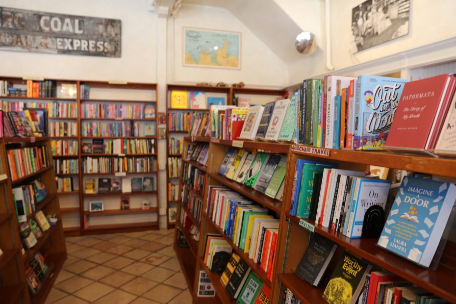 Bookshelves filled with various books line the walls and aisles of a cozy bookstore, with a tiled floor and decorative signs on the walls.