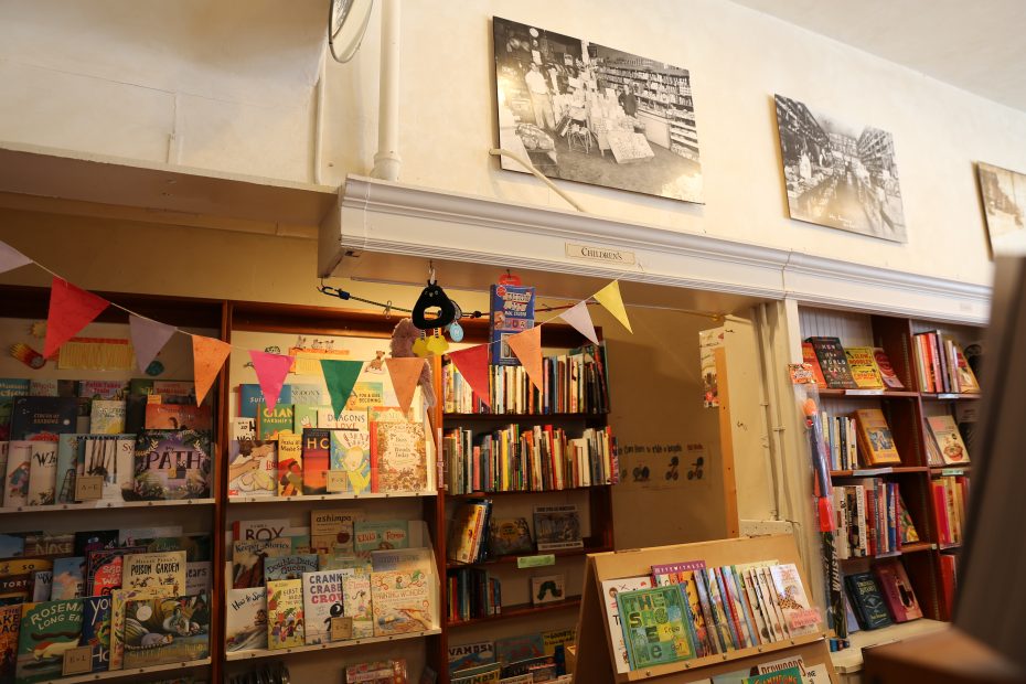 Children's section of a bookstore with shelves of books, colorful bunting, and black-and-white historical photos displayed on the wall above.