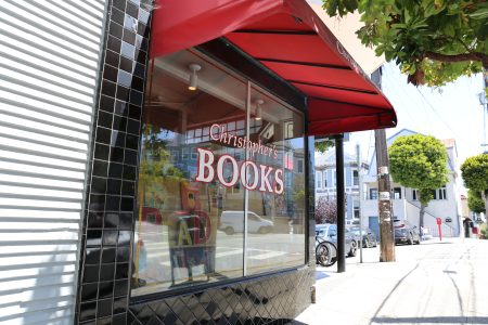 The storefront of a bookshop with a red awning and large window reading "Christopher's Books" on a sunny street corner.