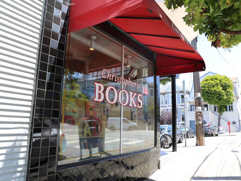 The storefront of a bookshop with a red awning and large window reading "Christopher's Books" on a sunny street corner.
