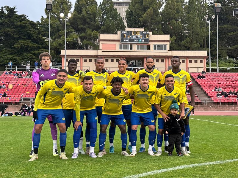 A soccer team in yellow jerseys poses for a group photo on a grass field, with a child in front and a stadium with red seats and scoreboard in the background.