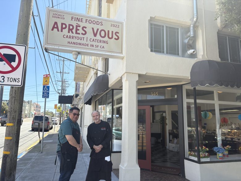 Two people stand outside the entrance of Après Vous, a carryout and catering shop in San Francisco, next to a sidewalk on a sunny day.