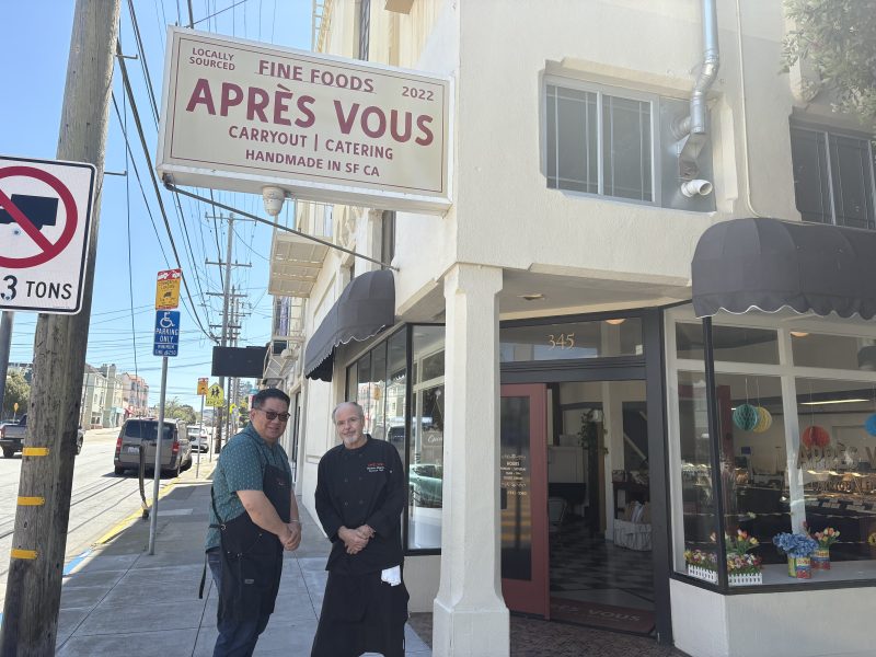 Two people stand outside the entrance of Après Vous, a carryout and catering shop in San Francisco, next to a sidewalk on a sunny day.