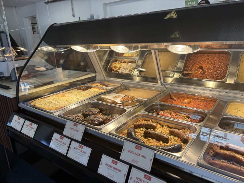 A deli counter displays various prepared foods in metal trays, including sausages, casseroles, and chili, with labeled signs in front of each dish.