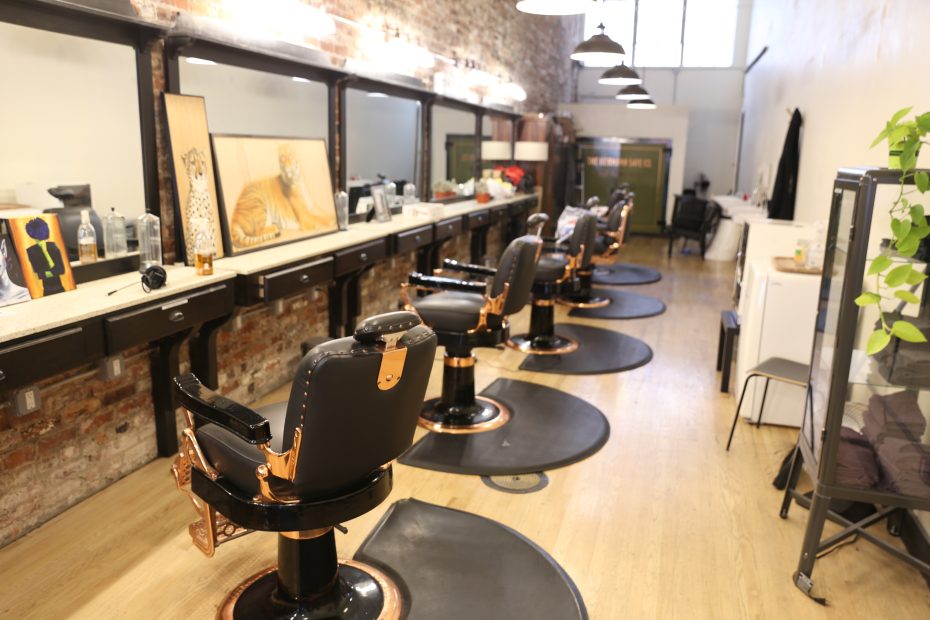 A row of empty black barber chairs faces mirrors along a brick wall in a modern, well-lit barbershop with wooden floors.