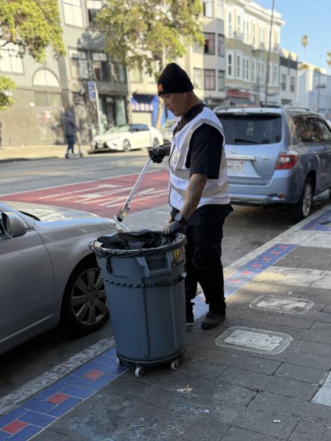 A person wearing a safety vest and beanie uses a grabber tool to pick up litter and place it into a large wheeled trash bin on a city sidewalk.