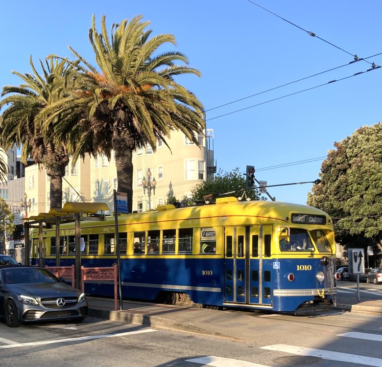 A yellow and blue streetcar labeled “1010” waits at a stop near palm trees on a sunny street, with a Mercedes parked nearby.