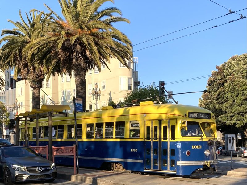 A yellow and blue streetcar labeled “1010” waits at a stop near palm trees on a sunny street, with a Mercedes parked nearby.