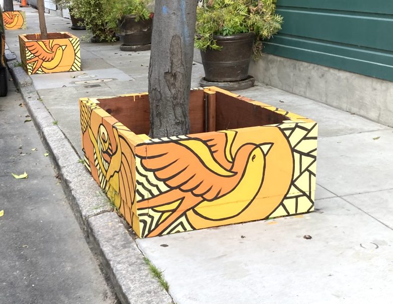 A tree on a sidewalk is surrounded by a wooden planter box painted with a large yellow and orange bird design.