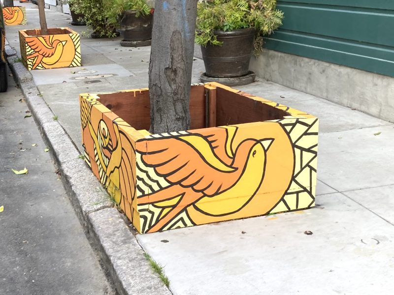 A tree on a sidewalk is surrounded by a wooden planter box painted with a large yellow and orange bird design.