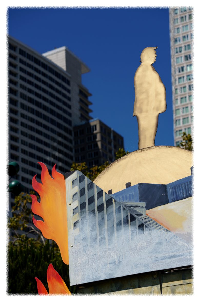 A cardboard cutout of a person stands atop a painted globe with flames and buildings, set against a backdrop of modern high-rise buildings under a clear blue sky.