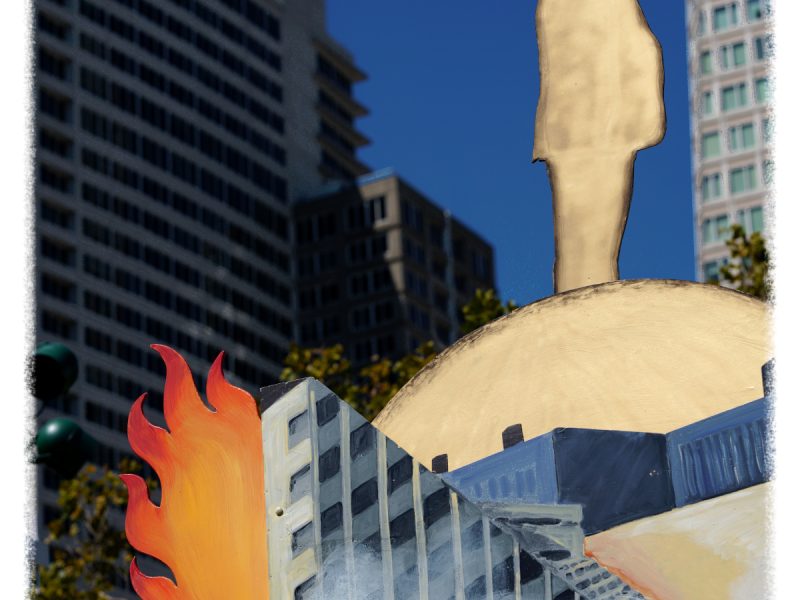 A cardboard cutout of a person stands atop a painted globe with flames and buildings, set against a backdrop of modern high-rise buildings under a clear blue sky.