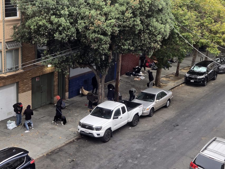 A group of people stand and sit on a sidewalk under trees next to parked cars and buildings on a city street.