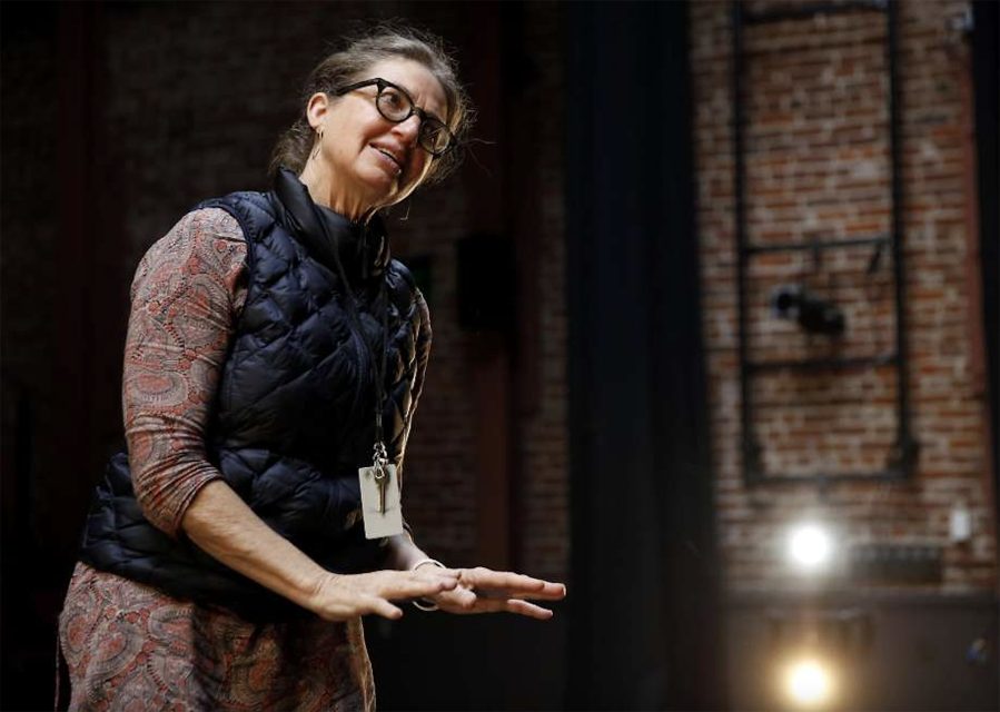 A woman wearing glasses, a patterned dress, and a black vest with an ID badge gestures with her hands indoors against a brick wall background.