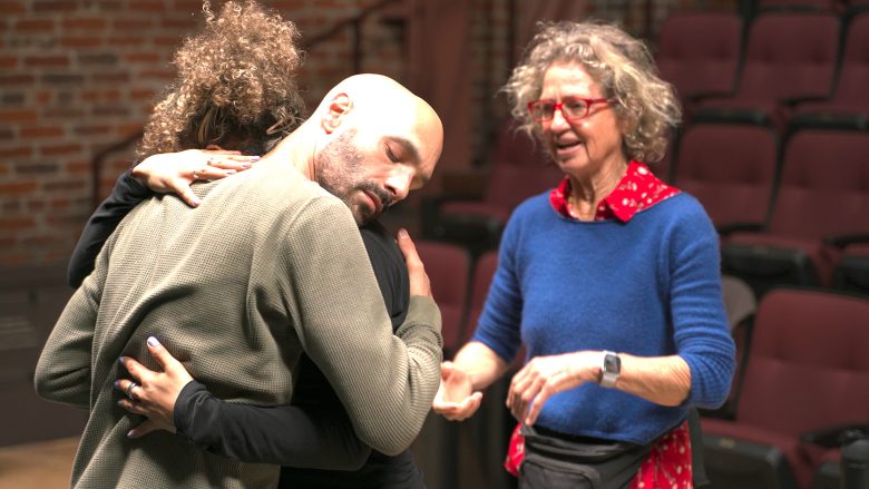 A man and woman embrace while another woman with curly hair, glasses, and a blue sweater stands nearby in what appears to be a theater setting.