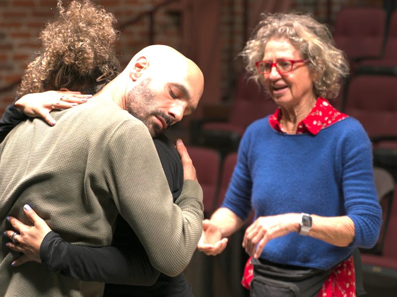 A man and woman embrace while another woman with curly hair, glasses, and a blue sweater stands nearby in what appears to be a theater setting.