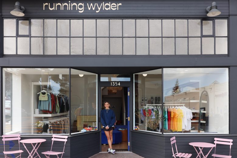 A person stands at the entrance of a store called "running wylder" with clothing and accessories displayed in the windows and pink tables and chairs outside.