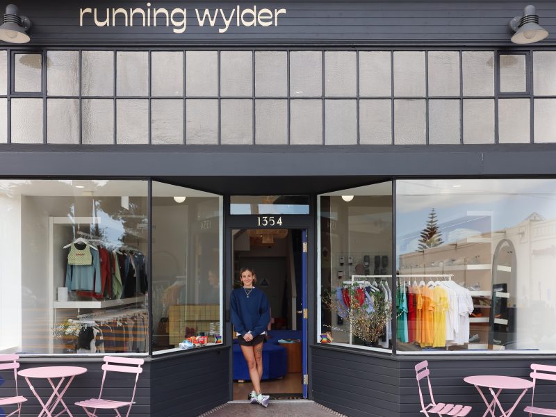 A person stands at the entrance of a store called "running wylder" with clothing and accessories displayed in the windows and pink tables and chairs outside.