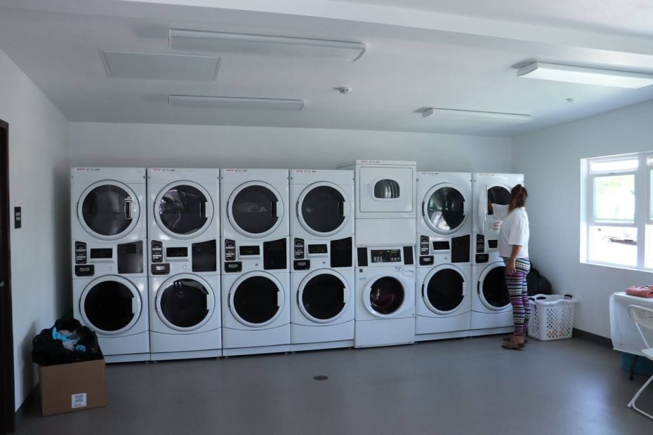 A woman loads clothes into a washing machine in a bright, clean laundromat with multiple washers and dryers lined against the wall.