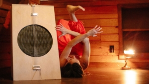 A person in a red outfit lies upside down with crossed arms near a large wooden box instrument in a dimly lit room with wooden walls and a lamp.