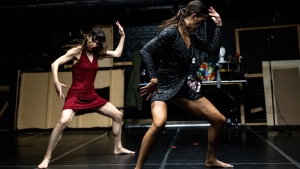 Two women perform a contemporary dance routine on a dark stage, each striking expressive poses with bent knees and raised arms.