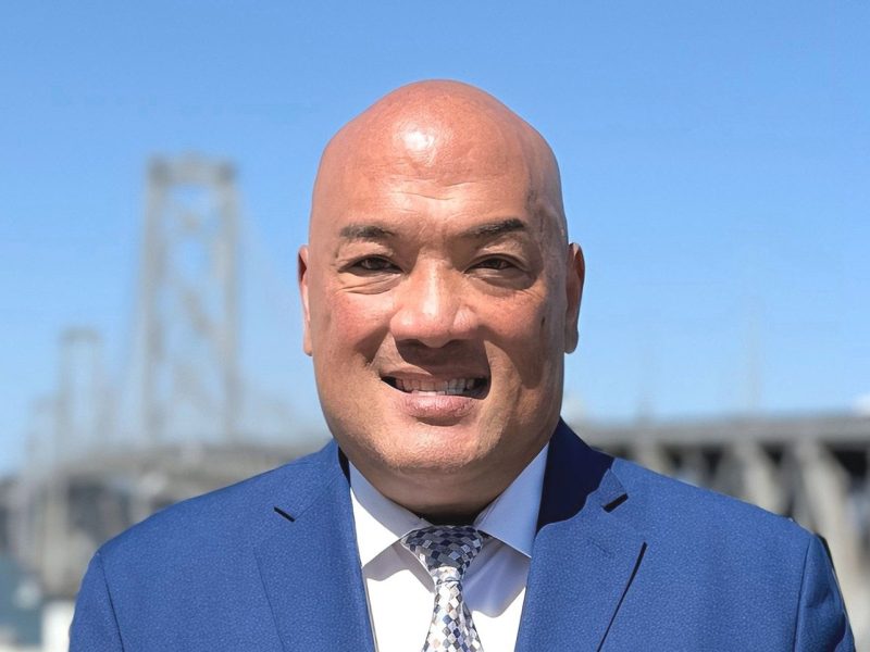 A bald man in a blue suit and patterned tie smiles outdoors with a large steel bridge visible in the background under a clear blue sky.