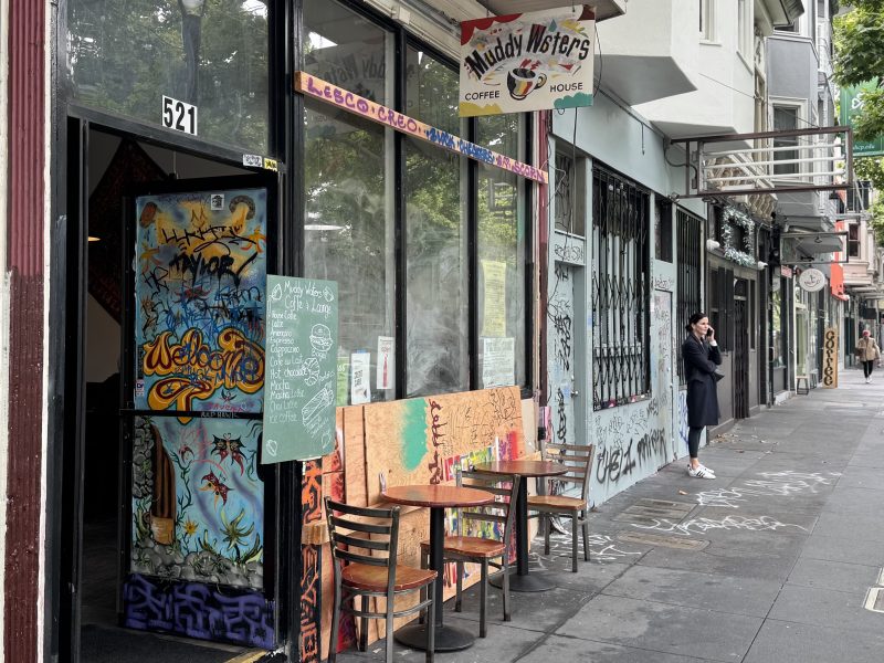Street view of Muddy Waters Coffee House with graffiti-covered door, outdoor tables and chairs, and a woman standing on the sidewalk using her phone.