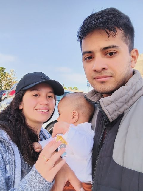 A man and a woman stand outdoors holding a baby. The woman is wearing a black cap and smiling, while the man looks at the camera. Cars and trees are visible in the background.
