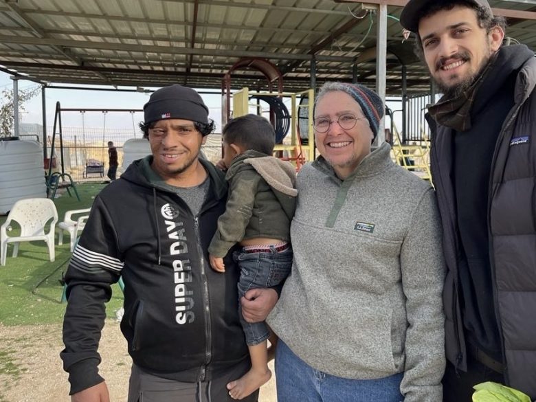 Four people, including a child held by an adult, stand together outdoors under a covered area, smiling at the camera. Some playground equipment and chairs are visible in the background.
