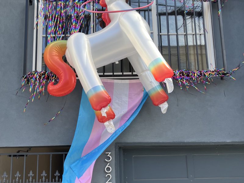 A large inflatable unicorn and a transgender pride flag hang from a second-floor balcony decorated with streamers above a building entrance.