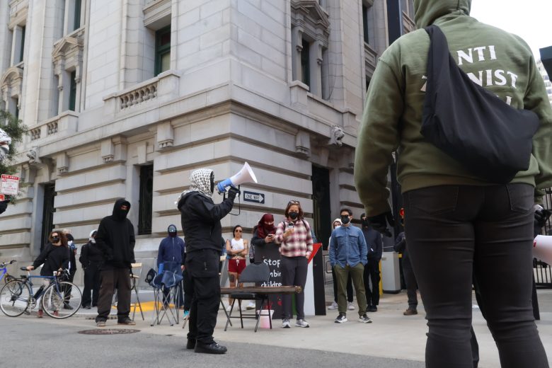 A group of people, some wearing masks and hoodies, gather outside a building. One person uses a megaphone while others hold signs. A bicycle is parked nearby.