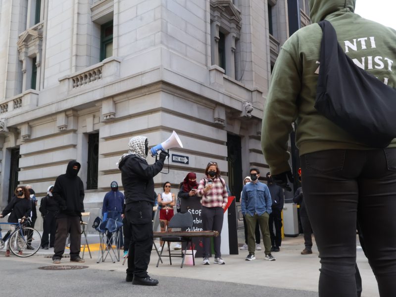 A group of people, some wearing masks and hoodies, gather outside a building. One person uses a megaphone while others hold signs. A bicycle is parked nearby.
