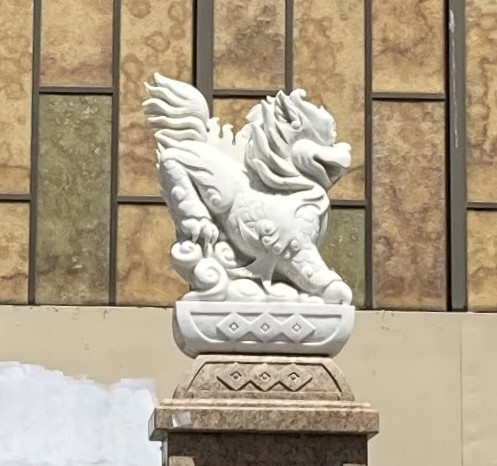 White stone statue of a mythical lion-like creature on a decorative pedestal, set against a tan and brown tiled building wall.