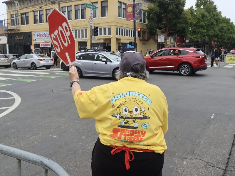 A volunteer wearing a yellow "Sunday Streets" shirt holds a stop sign at an intersection, directing traffic on a city street.