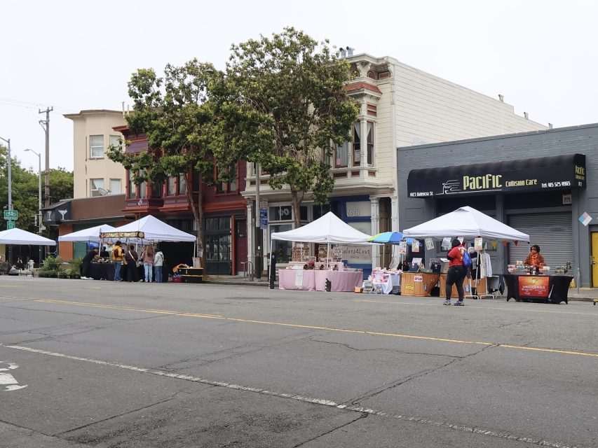 Outdoor street market with several vendor tents set up along the sidewalk in front of buildings, with a few people browsing stalls.