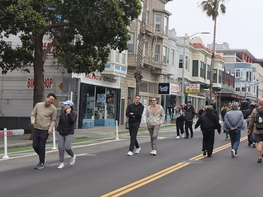 People walk along a city street lined with shops and trees, including a bike shop and a cycling store, on an overcast day.