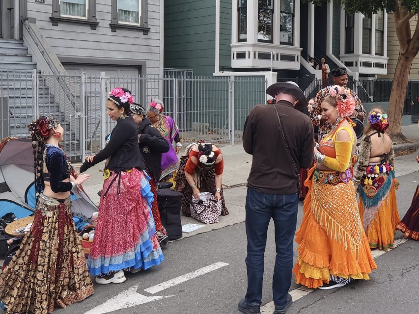 A group of people in colorful, traditional costumes stand and talk on a city street near Victorian-style houses. Some are adjusting outfits, and two people have cameras.