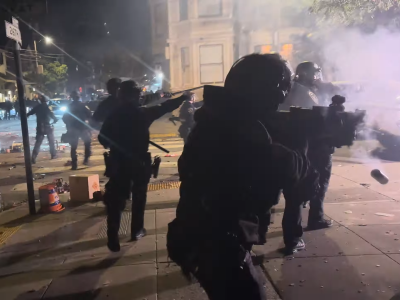 Police officers in riot gear deploy crowd control measures on a city street at night amid scattered debris and smoke.