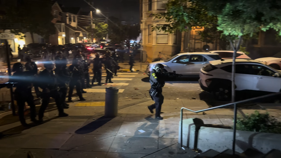 A police officer runs across a city street at night while others stand in formation nearby; damaged cars and debris are visible.