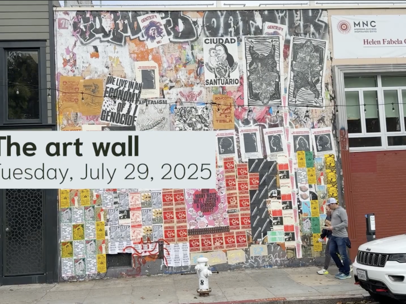 A wall covered with various posters, flyers, and street art, with two people and a dog passing by; text overlay reads "The art wall - Tuesday, July 29, 2025.