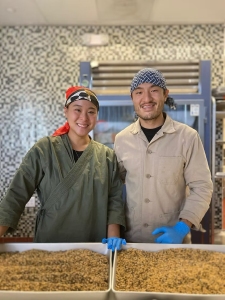 Two people in work attire and bandanas stand behind trays filled with grain or seeds, smiling in a kitchen or food preparation area.