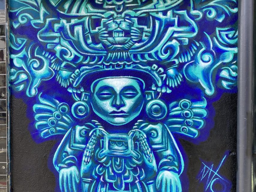 A mural depicts a stylized, blue-toned figure in traditional Mesoamerican attire, with an elaborate headdress and closed eyes, against a dark background.