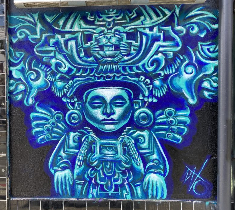 A vibrant mural depicts a stylized figure in elaborate, symmetrical headdress and attire, painted in shades of blue and turquoise with bold outlines.