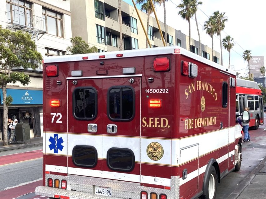 A San Francisco Fire Department ambulance marked "72" is stopped on a city street near buildings and palm trees.