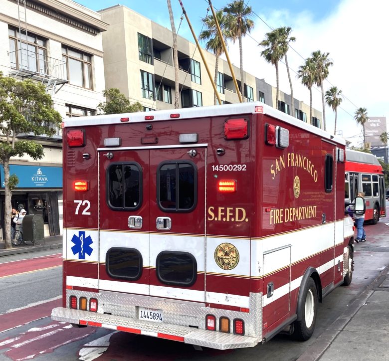 A San Francisco Fire Department ambulance is parked on a city street near buildings and a bus, with emergency lights visible but not activated.